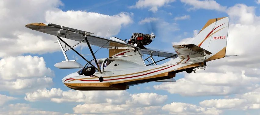 A Searey amphibious flying boat on its beaching gear beside the water, wing and pusher engine against a cloudy sky.